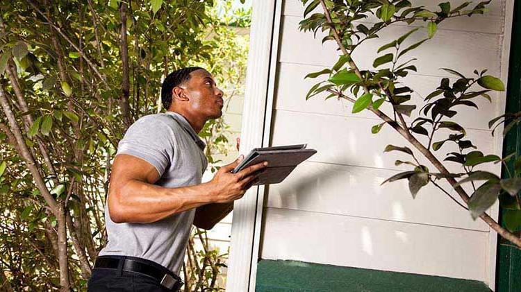 Man looking up holding a tablet while inspecting the exterior of a home.