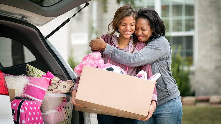 Mom hugging daughter holding box of dorm room supplies.